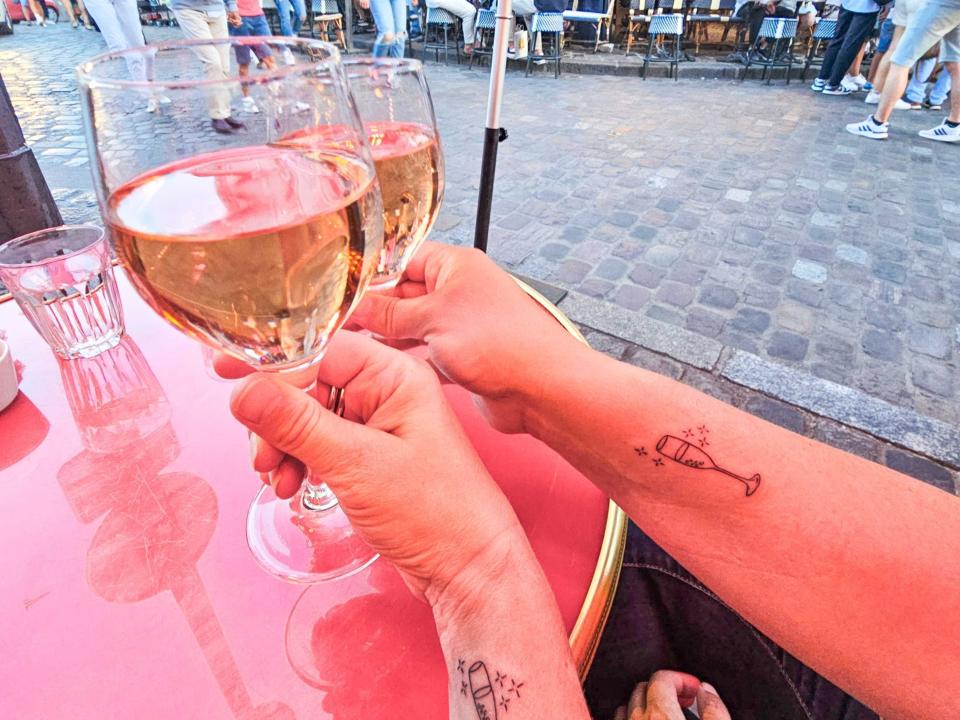 Two arms with Champagne flute tattoos clinking two wine glasses together over a red table. A cobblestone path filled with people is in the background