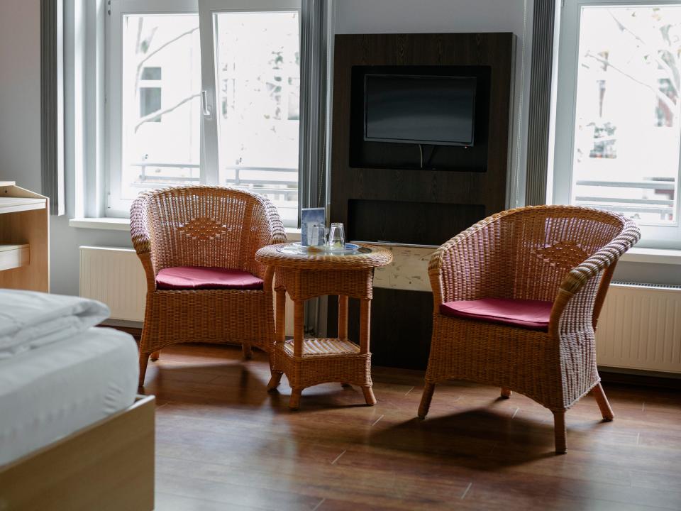 Bedroom with arched windows overlooking other building with rattan chairs in room with table in between and bed with white sheets in foreground