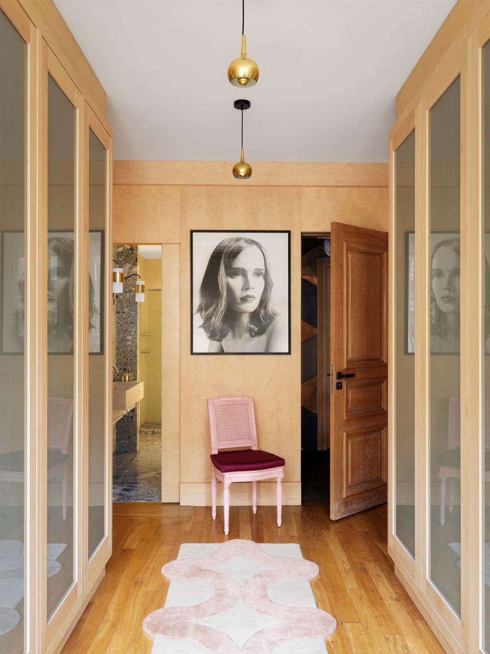 two sides of a walk in closet have three mirrored doors each, wood floors, small brass pendants, a pink chair with burgundy cushion, a closeup photo portrait of a young woman, and a carved wooden entry door