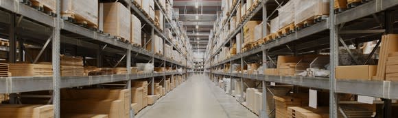 Aisle in warehouse with shelves containing pallets going up to the ceiling and bright overhead lights.