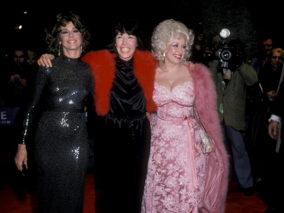 Jane Fonda in a black shiny gown, Lilly Tomlin in a black dress and red fur shawl, and Dolly Parton in a baby pink floral lace dress with a pink fur coat.