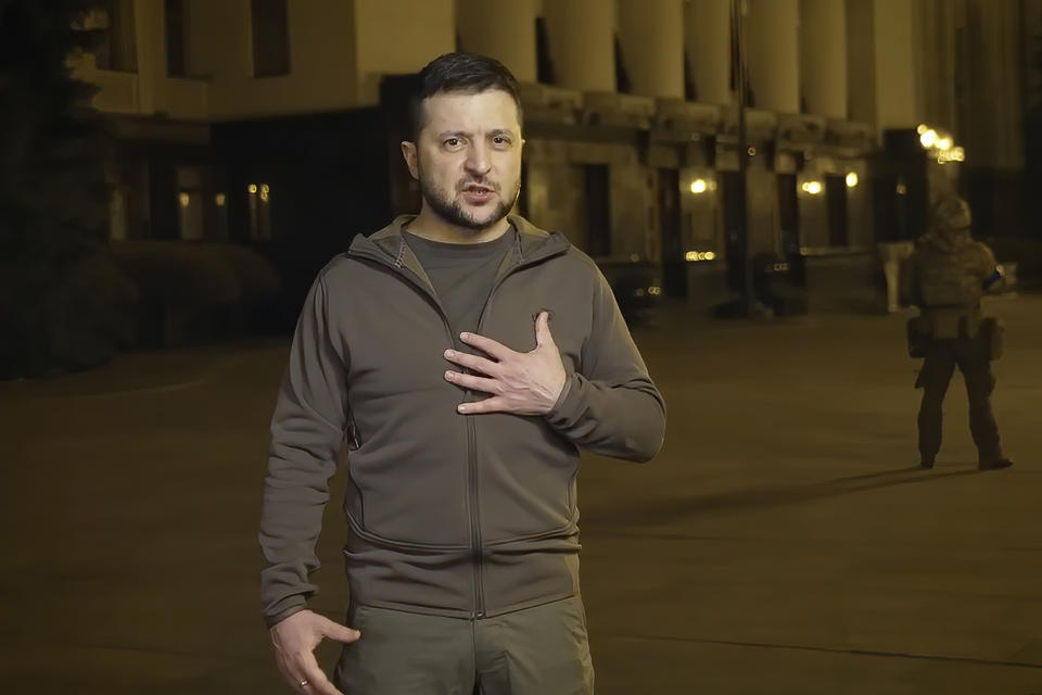 Volodymyr Zelensky holds his left hand over his heart while standing in front of a building with a colonnade near someone wearing a helmet and military gear.
