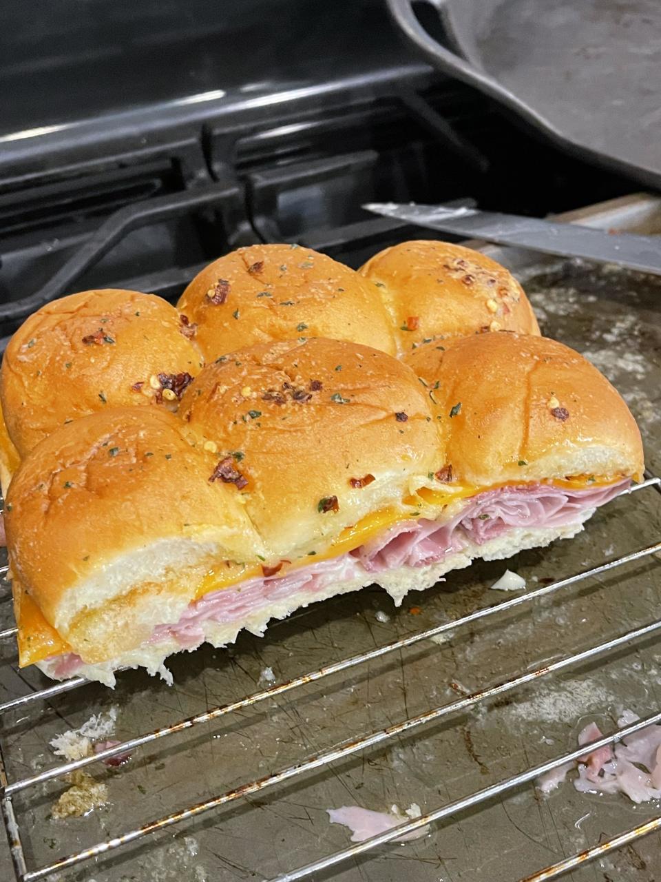 Six slider-sized sandwiches with ham and melted cheese, topped with a seasoned, slightly crispy bun, on a baking rack in a kitchen setting