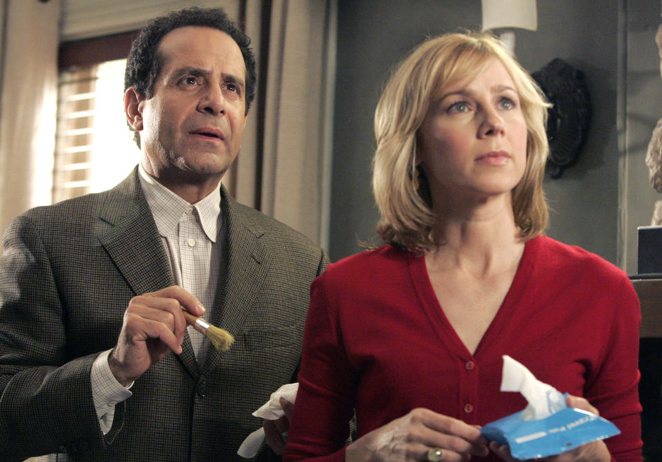 Two actors, one in a suit holding a small object and the other in a red top holding tissues, appear focused on something off-camera in a scene from the show "Monk."