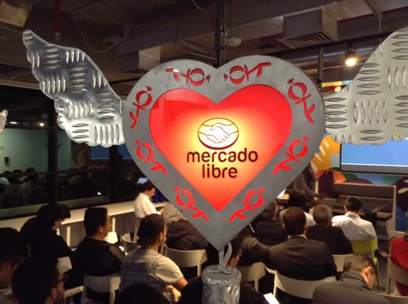 The MercadoLibre logo in a heart hovering over a crowd seated at a conference.