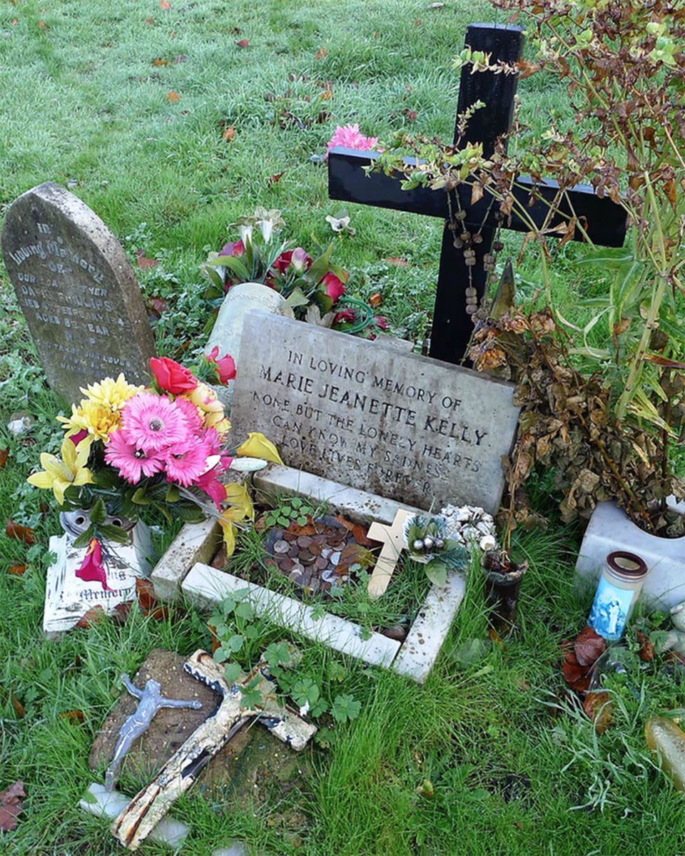 Mary Jane Kelly's grave in Leytonstone Roman Catholic Cemetery, London surrounded by several flowers, coins, and crucifixes with grass