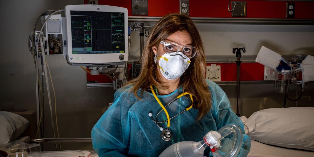 a female doctor in scrubs, goggles, and a mask looks directly into the camera in a hospital room
