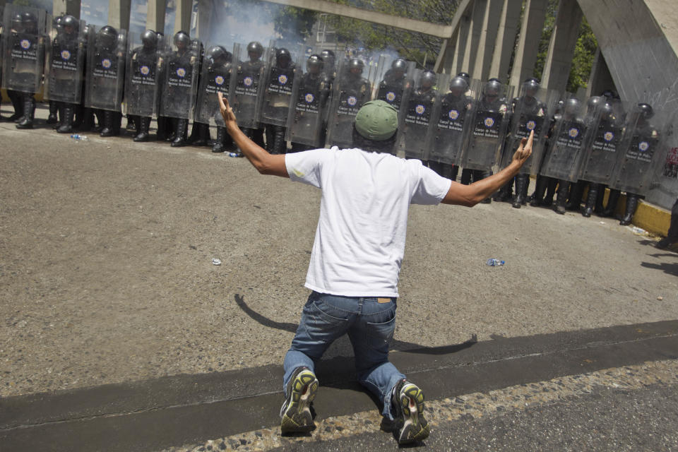 An anti-government demonsrator kneels in front of a formation of Bolivarian National Police in riot gear, during clashes at the Central University of Venezuela, UCV, in Caracas, Venezuela, Thursday, March 20, 2014. Thursday dawned with two more opposition politicians, San Cristobal Mayor Daniel Ceballos and San Diego Mayor Enzo Scarano, behind bars. Police used tear gas and water cannons to disperse a student-called protest of several thousand people in Caracas, some of those demonstrating against the arrests of the mayors. (AP Photo/Esteban Felix)