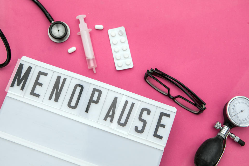 On a bright pink surface lie a sign saying 'menopause' in block letters, a stethoscope, syringe, capsules, blister pack, glasses and blood pressure gauge.