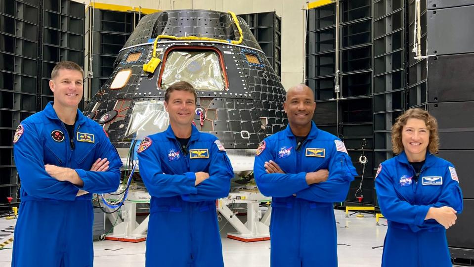 four astronauts standing in flight suits in a row. their arms are crossed. behind them is a cone-shaped spacecraft