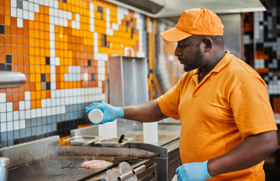 A man in an orange polo shirt and cap is grilling a burger patty, seasoning it in a professional kitchen. He is wearing blue gloves for hygiene