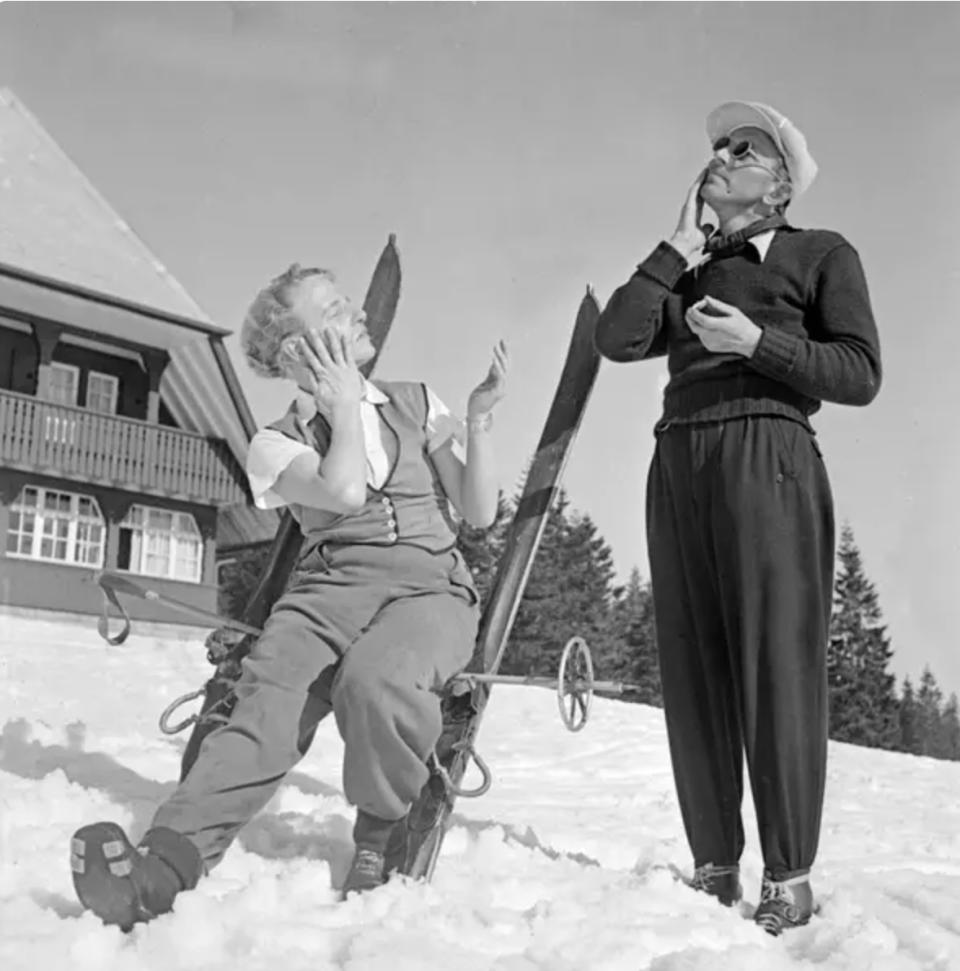 Two people in winter clothing enjoying a snowy day; one is sitting on skis, the other standing nearby, both covering their eyes from the sun