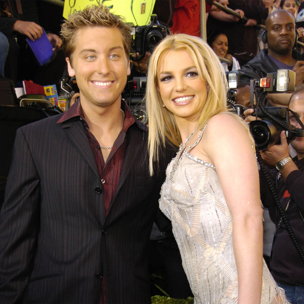 Lance Bass and Britney Spears smile on a red carpet. Bass is in a black suit with a red button-down shirt and Spears is in a sparkly, white gown (KMazur / WireImage)