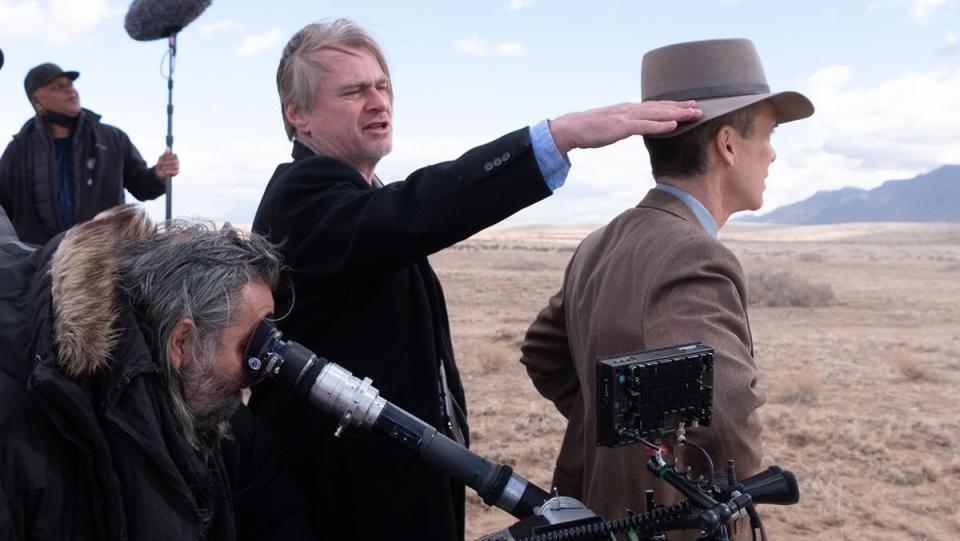Christopher Nolan's hair is blown by the wind while directing and surrounded by cast and crew on Oppenheimer