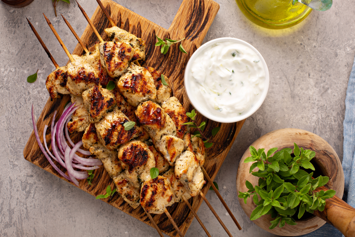 Top-view of five skewers of Easy Chicken Souvlaki on a wooden serving board with a white bowl of Tzatziki and onions, olive oil and herbs on the right, on a granite table