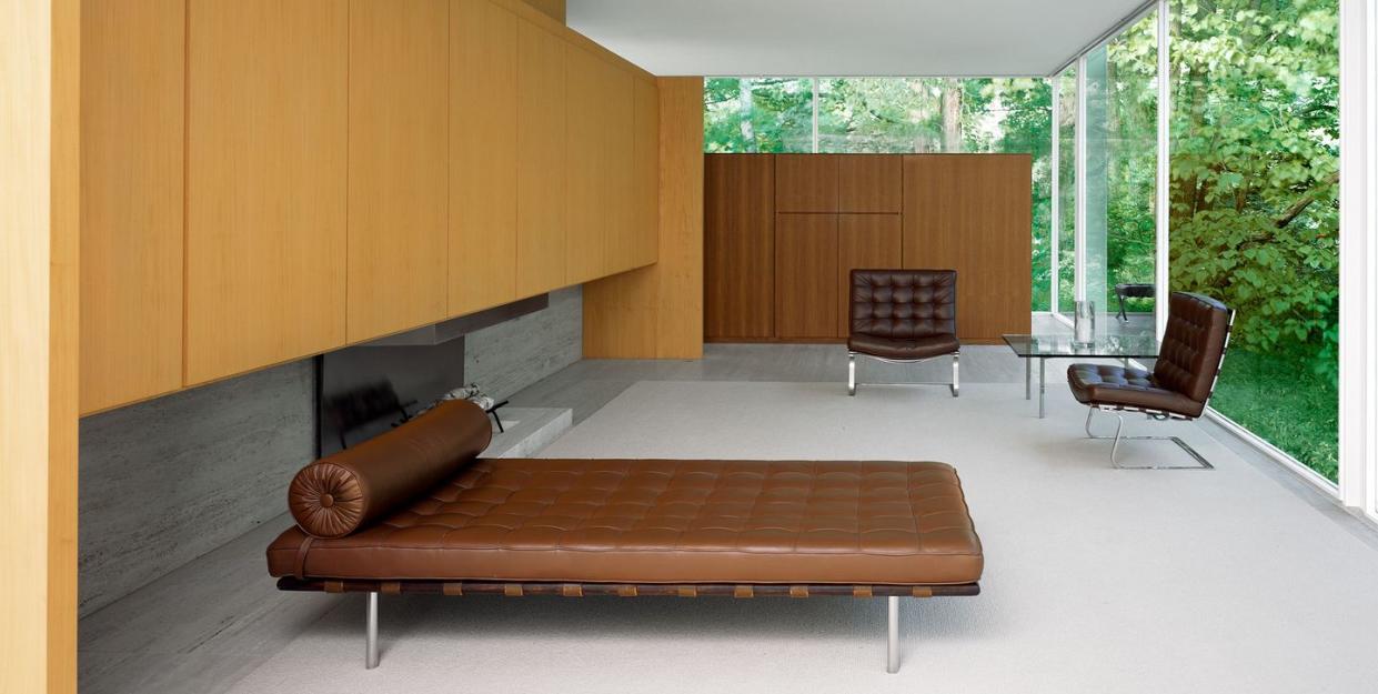 a living room with glass walls on two sides looking out onto leafy green trees, furnished with a brown leather daybed and two brown leather barcelona chairs and light wood cabinets against a long wall