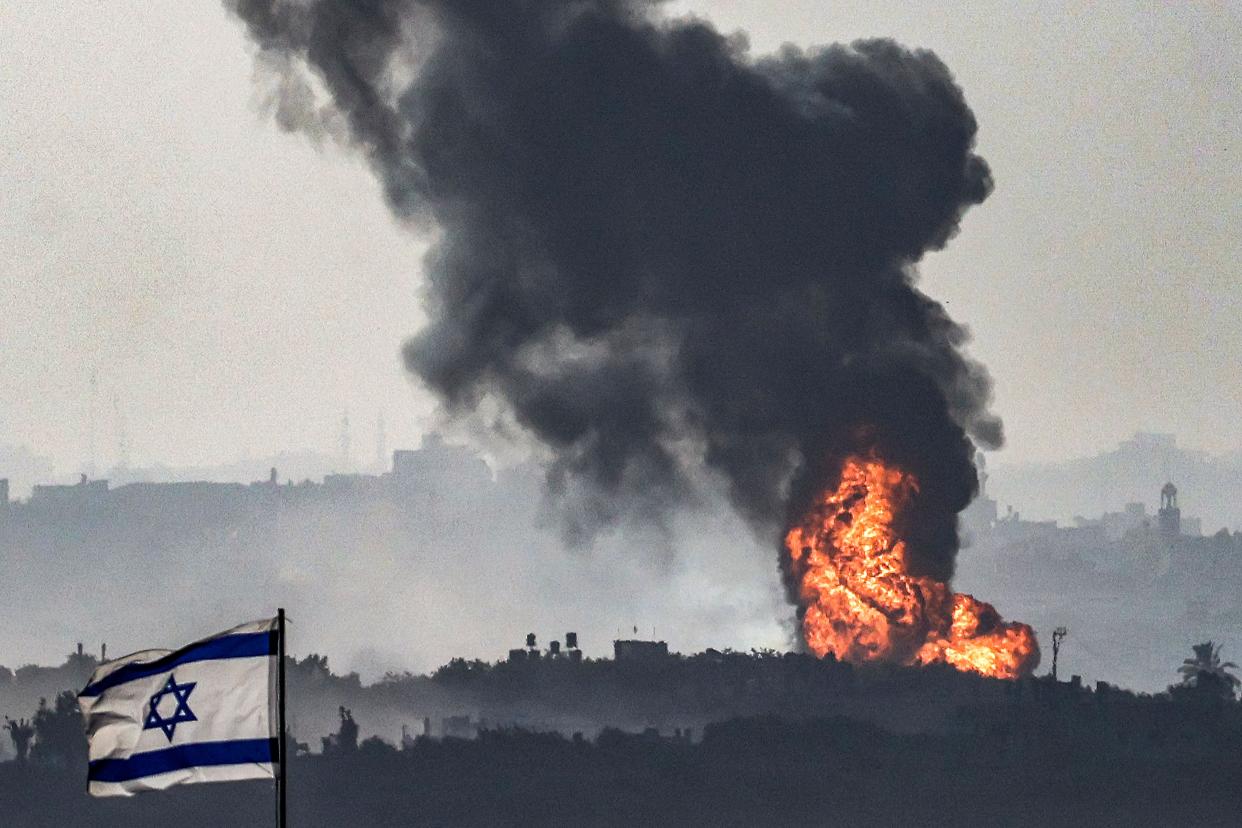 An Israeli flag in the foreground while a fireball erupts and releases thick smoke in the background.