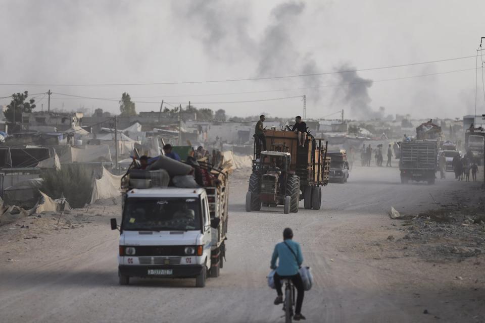 Several people in vans and a tractor on a dusty street with smoke rising in the background.