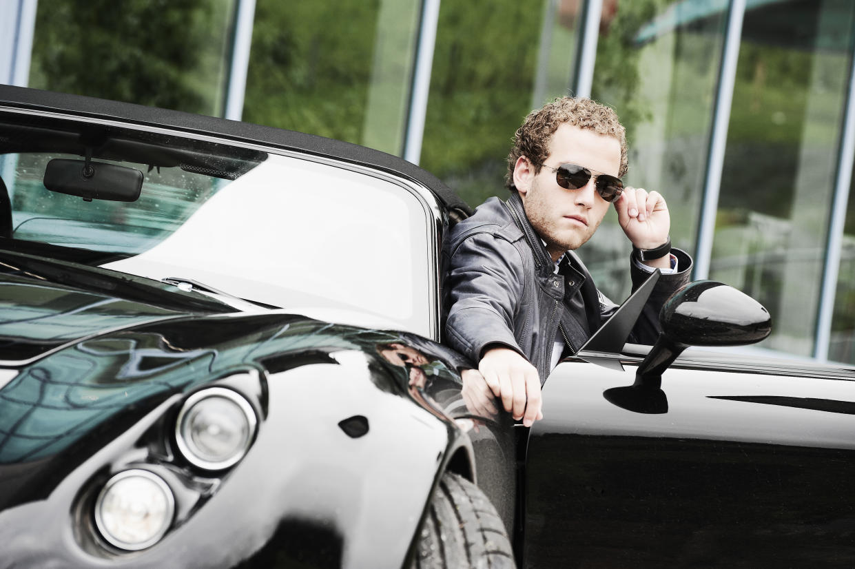 A young, male entrepreneur wearing sunglasses poses while sitting in a sportscar.