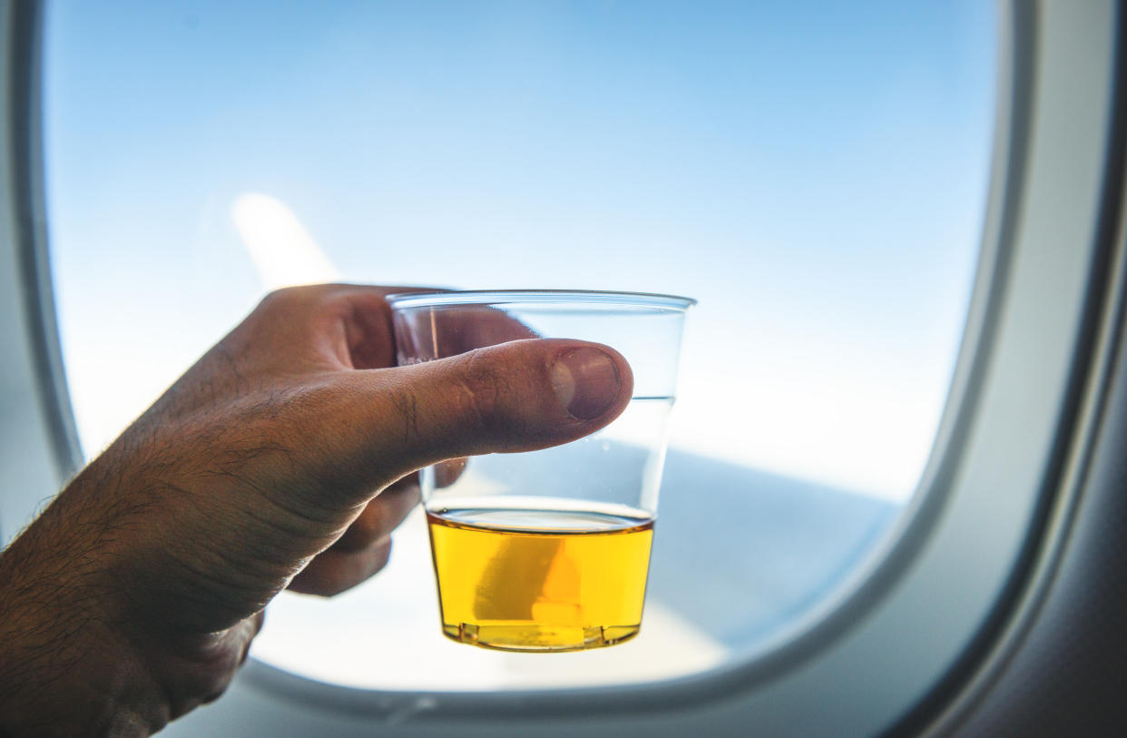 A hand holds an alcoholic beverage in a plastic cup next to an airplane window.