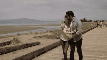 A couple is walking arm-in-arm on a boardwalk near a scenic beach with mountains in the background. The man wears a button-up shirt and jeans, the woman wears a t-shirt and jeans