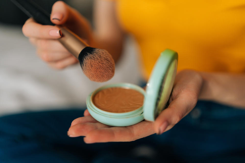 One hand holds an open makeup compact while the other holds a makeup brush poised to swipe across the makeup