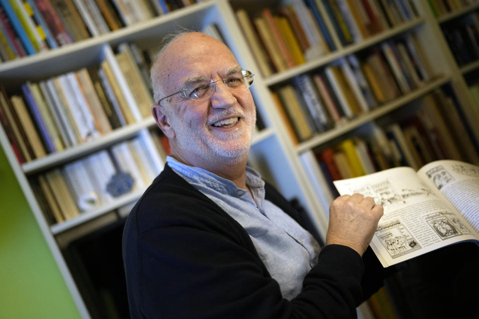 Osvaldo Sousa laughs while showing one of the cartoon books he has published, during an interview in his apartment in the suburbs of Lisbon, Feb. 28, 2024. Sousa, an opera singer at Lisbon's Sao Carlos theater who as a 20-year-old student witnessed first-hand tanks and troops in the streets of Lisbon on April 25, 1974, says "Our dreams came up short". (AP Photo/Armando Franca)