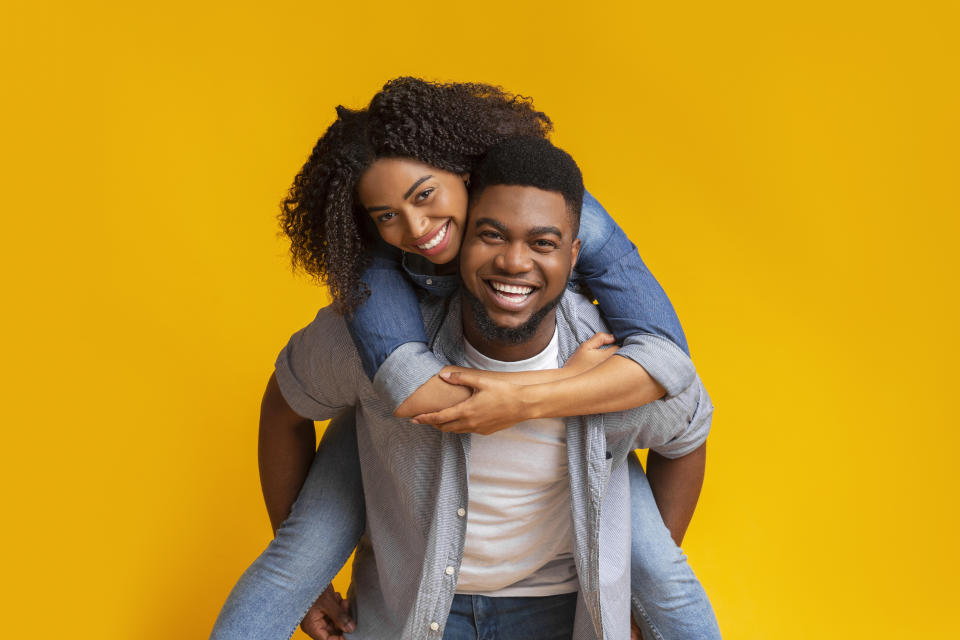 Piggyback Ride. Cheerful Black Boyfriend Piggybacking His Happy Girlfriend, Couple Fooling Together Over Yellow Background In Studio, Free space
