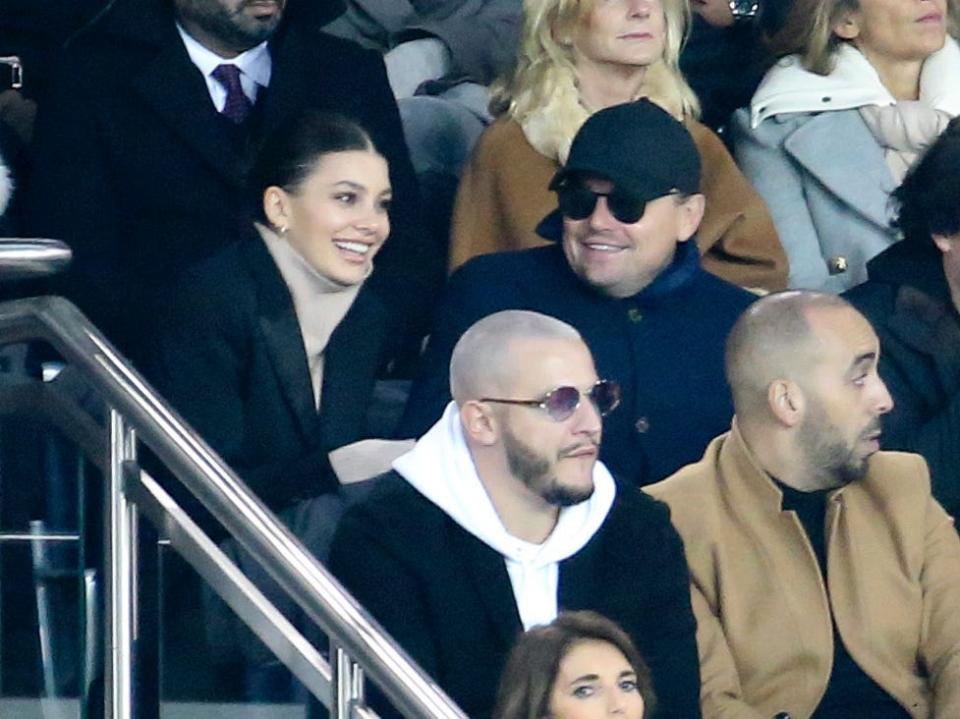 Camila Morrone, in a dark jacket, and Leonardo DiCaprio, in a black baseball hat and dark sugnlasses, smile at each other while sitting in the crowd at a soccer game in Paris.