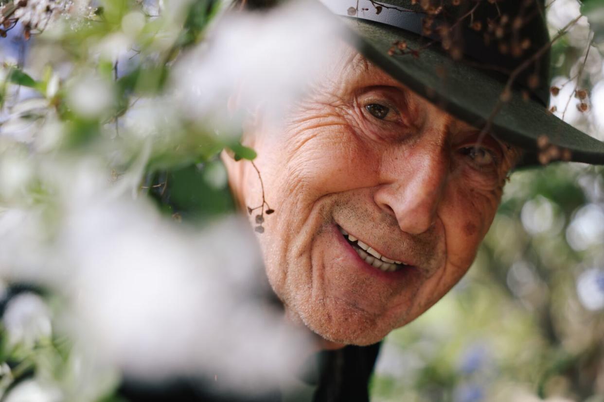 A smiling man in a hat peers through some garden plants