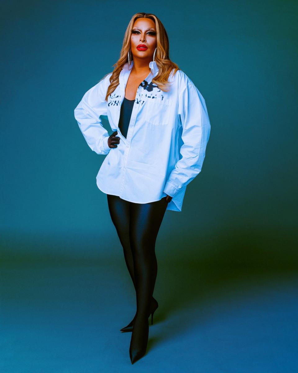 Roxxxy Andrews in a loose white button-down shirt, black tights, and high heels posing confidently against a plain background