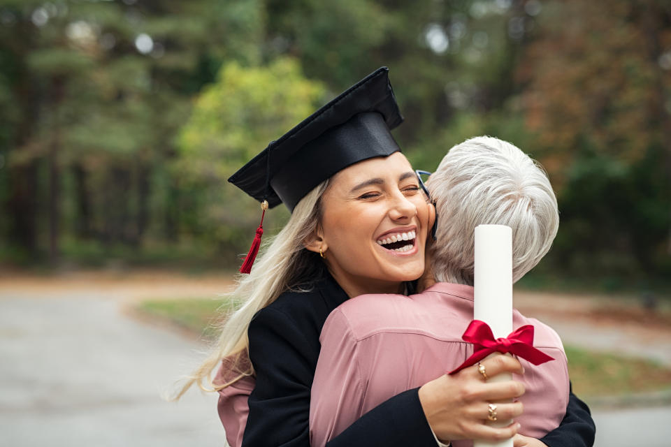 Enthusiastic graduated daughter holding degree embracing mother in campus. Young female student graduate hugging her mother at graduation ceremony. Excited college student with the graduation gown and hat holding diploma and hug the parent at campus.