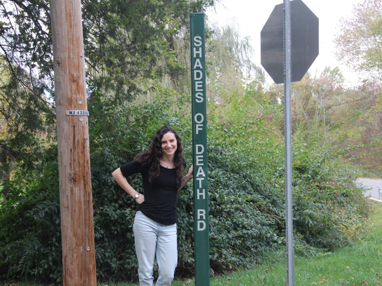 A woman stands next to a road sign reading "Shades of Death Road."