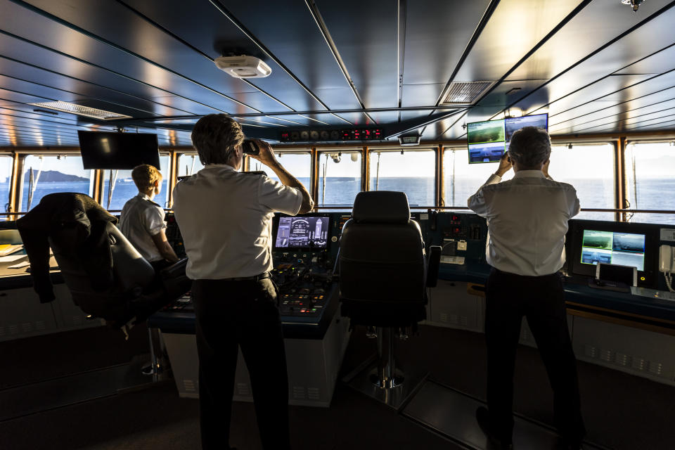 Three people in uniforms are working on the bridge of a ship, using various instruments and monitoring screens, with an ocean view in the background