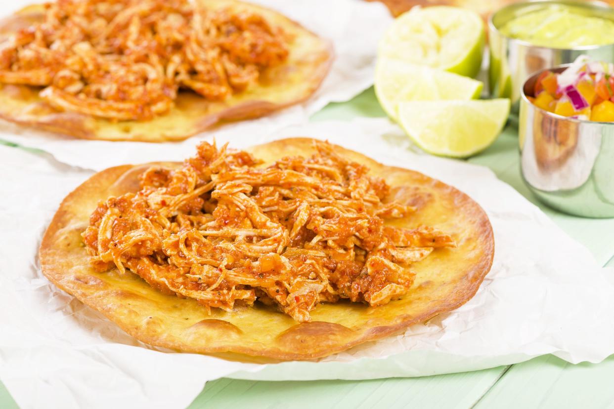 Two mango-chicken tinga tacos on white serving papers on a seafoam wooden table with a background of ingredients