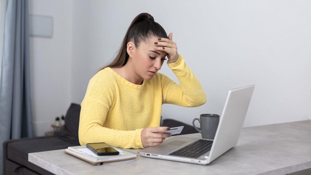  A white woman with long brown hair in a ponytail looks down at her computer in a distressed manner. She is holding her forehead with one hand and a credit card with the other. 