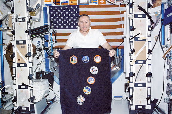 On board the International Space Station in 2002, astronaut Jerry Ross displays the mission logos representing the record seven space shuttle missions he has flown