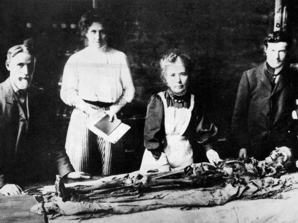 A black and white picture shows four Victorian egyptologists unwrapping a mummy