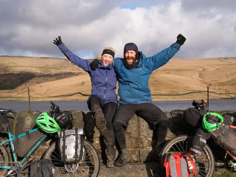 Joshua Kian and Sarah Morgan sitting on a wall near two bikes with their hands in the air.