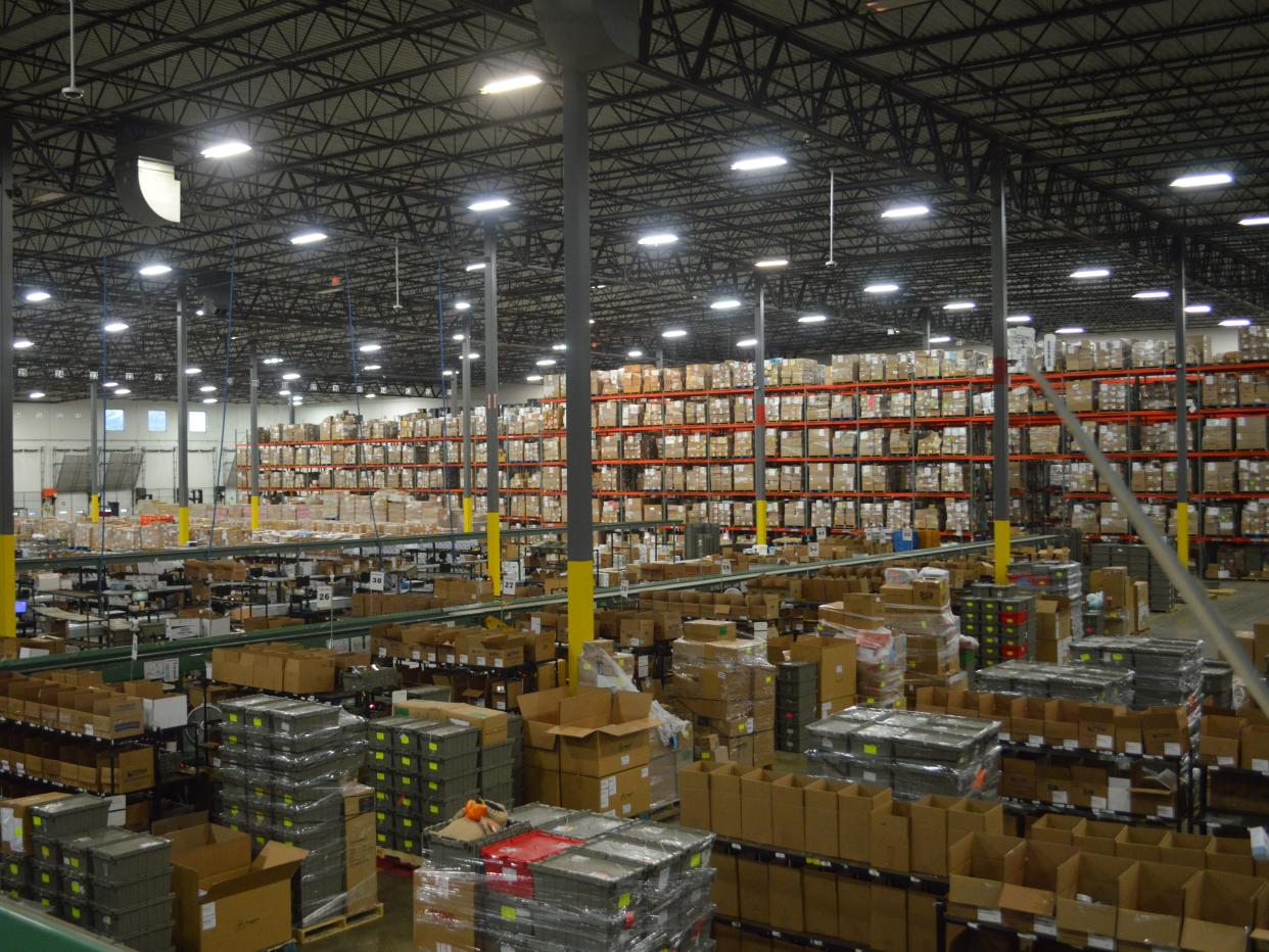 Cardboard boxes on pallets are stacked six levels high on orange shelves in a gray warehouse with overhead lighting.