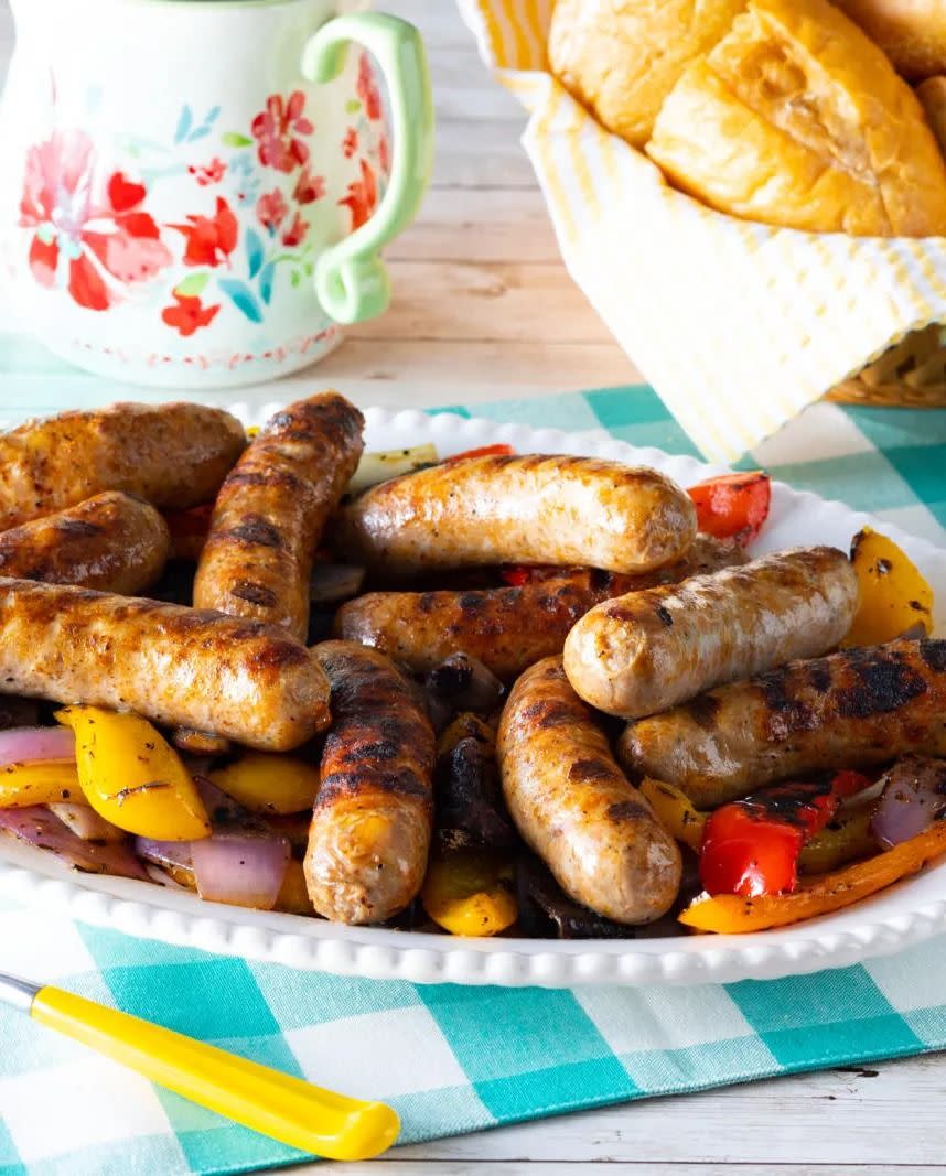 grilled sausages peppers on white platter with rolls in back