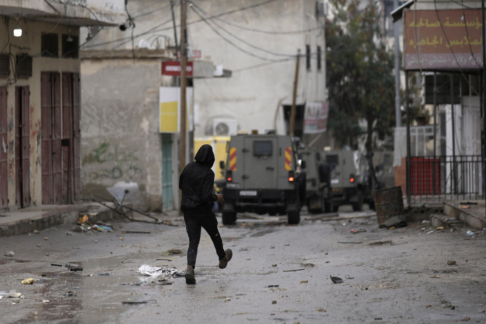 A Palestinian throws rocks at Israeli military vehicles raiding Aqbat Jabr camp, southwest of the city of Jericho on Saturday, Feb. 4, 2023, during a search for the Palestinian suspects behind a shooting attack at a restaurant in a nearby settlement last week. The army said it surrounded several residences that Palestinians were using to hide out and plan future attacks and ultimately arrested several suspects who surrendered. During the raid, the army said Palestinian protesters shot at Israeli security forces, who responded with live fire. The Palestinian Health Ministry said six people were wounded, two seriously. (AP Photo/ Majdi Mohammed)