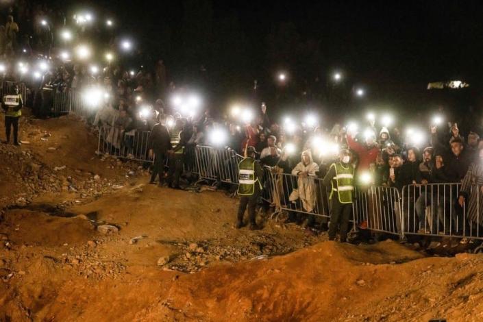 People stand behind a cordon. They use their mobile phones as torches.