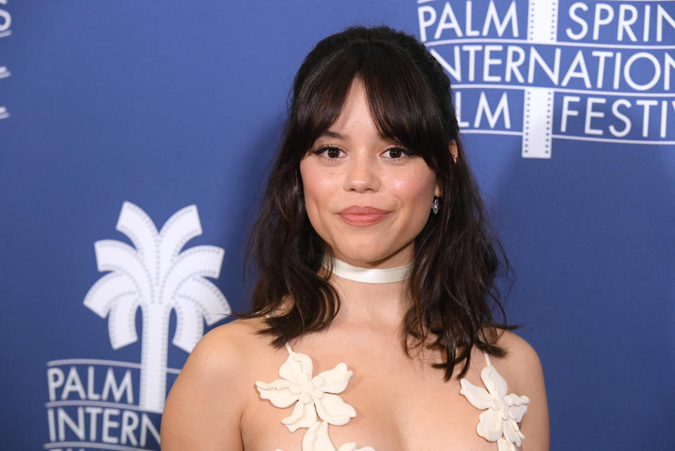 Jenna Ortega at the Palm Springs International Film Festival, wearing a floral white dress and a choker necklace, posing in front of the event's backdrop