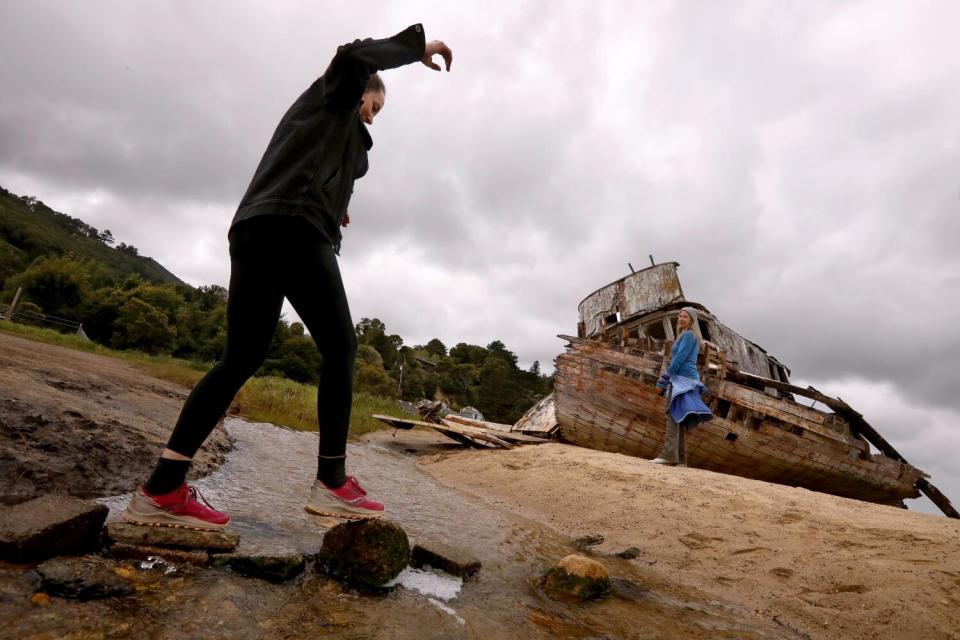 A woman walks across a rocky shoreline near an abandoned fishing boat.