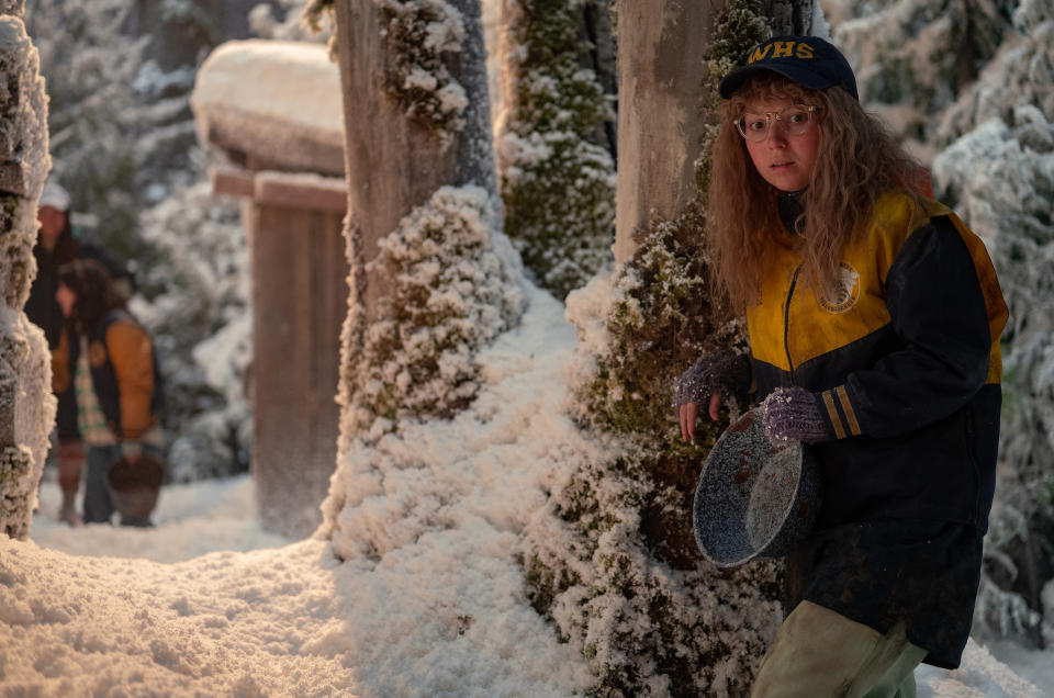 A teen girl in a cap and yellow and blue athletic jacket stands by a tree in the snowy wilderness, listening to others in the background; still from "Yellowjackets"