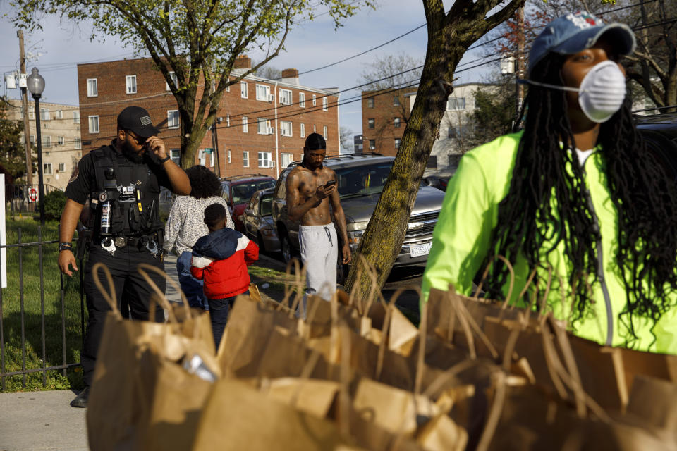 In this March 29, 2020, photo, people walk by an apartment complex security guard, at left, as entrepreneur and community volunteer India Blocker-Ford, right, stands by bags of donated hot food as she waits for members of the community to line up, in southeast Washington. Neighborhood deliveries are part of a new Martha's Table initiative, along with community partners, to get needed food directly to the neighborhoods they serve. (AP Photo/Jacquelyn Martin)