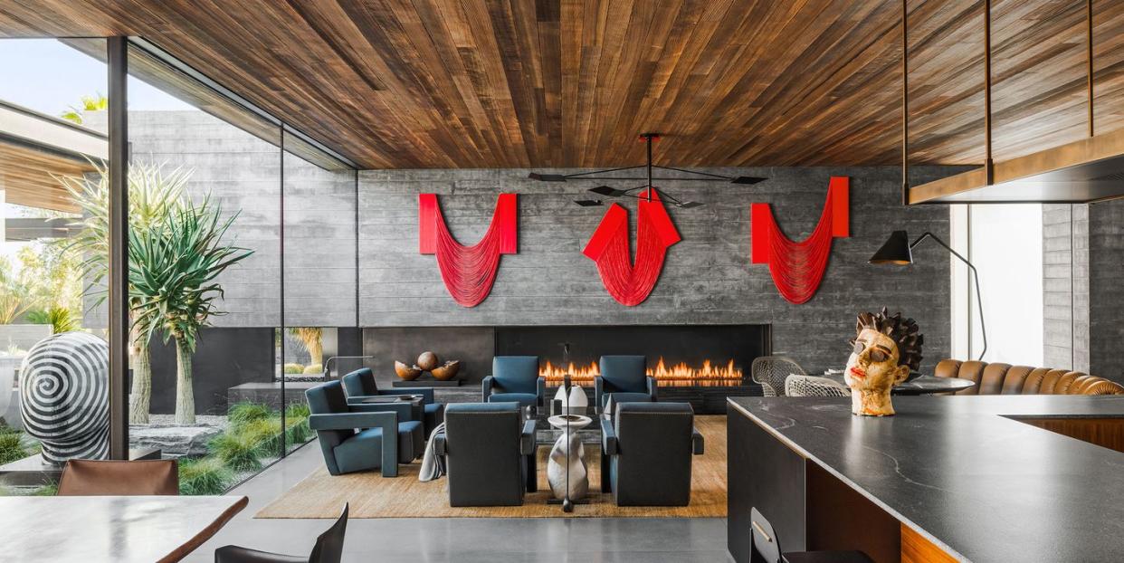 open kitchen dining living area with long black counter and stools and a gray brick wall with modern fireplace with fire in it and three large red pieces of artwork above it and a wood panelled ceiling and open glass walls on the left