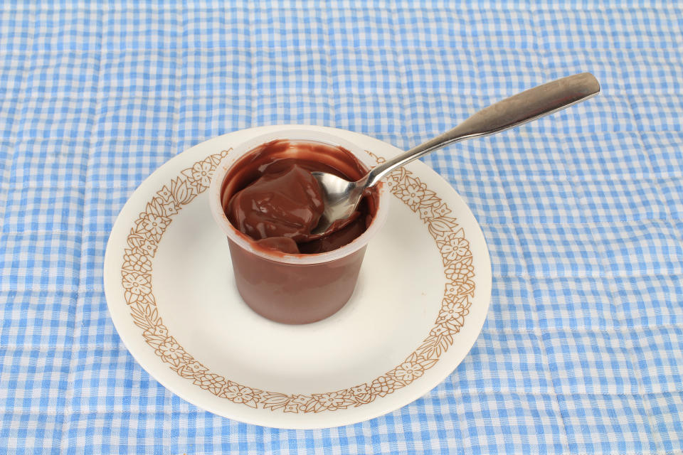 A spoon is placed inside a cup of pudding on a white plate with an ornate border, set on a checkered tablecloth