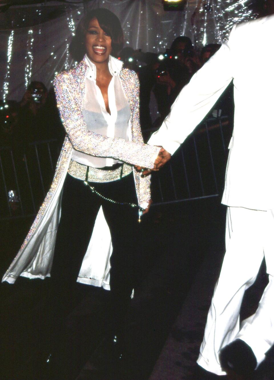 Whitney Houston walks down the red carpet at the 1999 Met Gala, grasping the hand of a man in a white suit. Houston is wearing black pants, a sheer white top, and sparkling duster jacket.